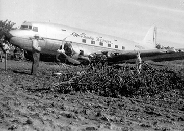 Monochromatic photograph shows a passenger aircraft resting on its fuselage. The left engine propellors are bent. A man and boy are standing next to a fallen tree in the foreground.