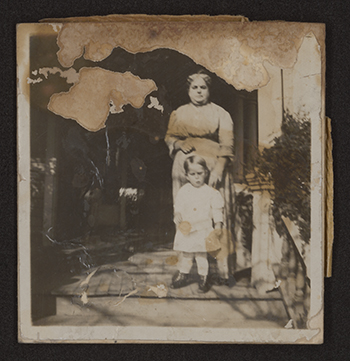 A monochromatic snapshot with surface damage and remnants of adhesive pressure senstive tape shows an older woman standing on the porch of a home. A young boy is standing in front of her.