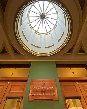 Image shows a vertical view looking up at a dome skylight. The circular center window has 16 spokes radiating outwards. The walls of the dome have vertical, rectangular moldings.  There is a wooden plaque on the green wall below the skylight with a carving of the Tarrant County Courthouse. The plaque was made by inmates from the Texas Department of Criminal Justice (TDCJ). The upper parts of the two sets of double doors are visible on either side of the green wall.
