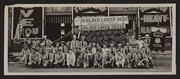 Monochromatic panoramic photograph of a large group of people, mostly men, posed in front of a delivery truck loaded with beer barrels and a sign on top that reads Drink Golden Lager Beer, 100 percent Union Made and Delivered, Golden Lager Beer.  A man at the back left of the group is holding a United States flag. A pole with a fabric sign at the right front of the group reads Brewery Workers Union No. 108 Fort Worth Texas. Walter Bob Berger, Sr. is the man standing next to the seated middle group and is wearing a dark tie and light cap.