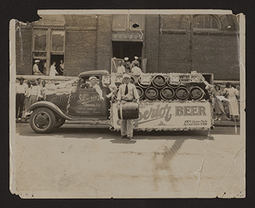 Monochromatic photograph of a man, Walter Bob Berger Sr., standing in front of a delivery truck loaded with beer barrels. The man is holding a beer barrel and has a cigarette in his mouth. The truck has text on the driver's side door for Superior Brewing Co. and a banner on the side reads Superior  Beer. The truck is decorated for a parade. Men and women, including some men of color, are standing behind the truck in front of a brick building.