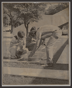 Black and white photo of an older man leaning down to point at a painting which is lying on the ground outdoors where a younger man is painting it.