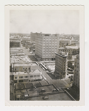 Monochromatic photograph with overall light brown tone shows an elevated or bird's eye view of a downtown street scene. Other buildings of varying sizes are visible.