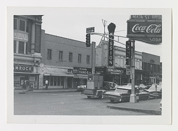 Black and white photograph shows a busy intersection with traffic. Different business storefronts, along with signs, are visible. A sign for Main Street Drug Store, advertising Coca Cola, is visible above a traffic light at the top right.