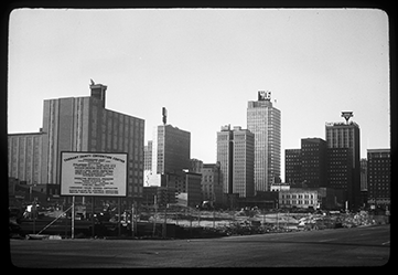 Black and white image shows a fenced off construction site with materials and equipment. A sign at the lower left foreground is headed with Tarrant County Convention Center and contains decision makers and vendors involved in the construction. Multiple tall office buildings are visible in the background.