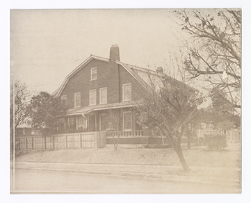 Monochromatic photograph with overall light brown tone shows an angled side view of a two and a half story brick house. The image prespective emphasizes the gambrel roof.