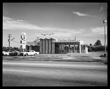 Black and white photograph shows a street in the foreground. There is a Mid-Century Modernist style building in the center with stone facing and zigzag canopies.
