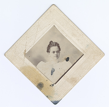 Monochromatic mounted photograph with diamond orientation shows a young woman from the lower chest up. She is wearing her hair in a topknot, glasses, and a light colored blouse with leg of mutton sleeves. There are multiple blue ink stains on the image and mount.