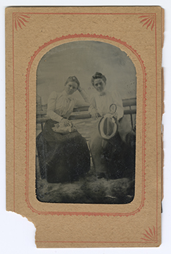 Monochromatic tintype photograph mounted in a tan paper frame with decorative red border. Full length image shows Annie, left, and Frances, right sitting on a bench in front of a painted backdrop. Both women are wearing high collared blouses tied with bows and holding hats in their hands.