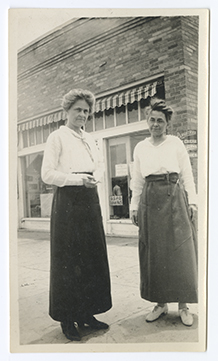 Full length monochromatic image with overall light brown tone shows two women standing on a sidewalk in front of a brick building.
