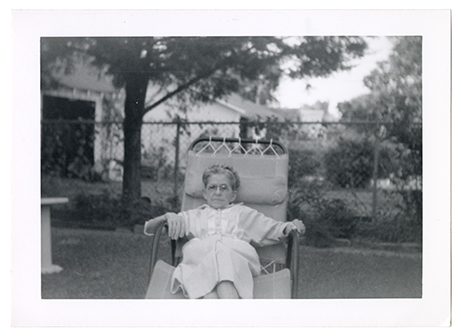 Monochromatic image shows a very small, elderly woman reclining in a sun lounger in a garden or yard. She is wearing glasses and both of her hands are resting on the arms of the chair.