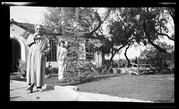 Monochromatic image shows an elderly man and woman standing in front of a Spanish style home. The man, left, is wearing a striped robe and holding a cigarette up to his mouth. 