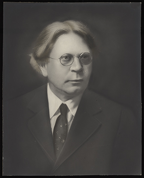 Monochromatic photograph of an older man from the chest up. He is wearing a dark suit and tie with a white shirt and round metal glasses.